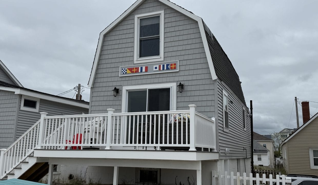 Gray shingled beach house with a white railing, small deck, and colorful nautical flags above the door, set on sandy ground near neighboring houses.