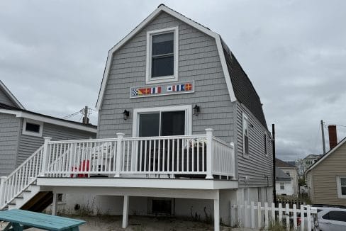 Gray shingled beach house with a white railing, small deck, and colorful nautical flags above the door, set on sandy ground near neighboring houses.