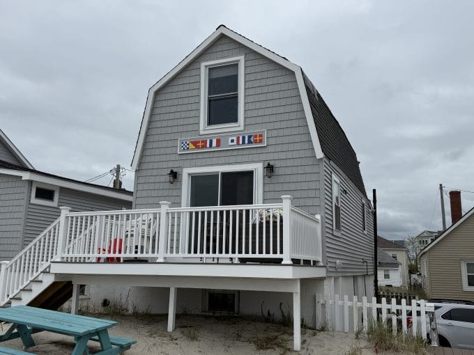 Gray shingled beach house with a white railing, small deck, and colorful nautical flags above the door, set on sandy ground near neighboring houses.
