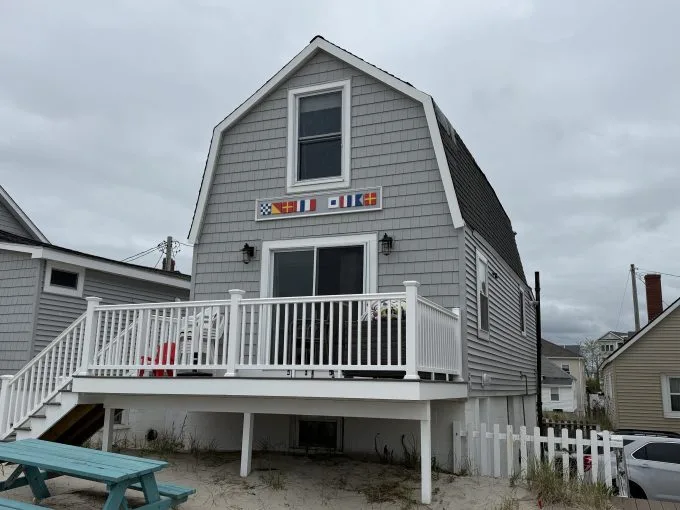 Gray shingled beach house with a white railing, small deck, and colorful nautical flags above the door, set on sandy ground near neighboring houses.