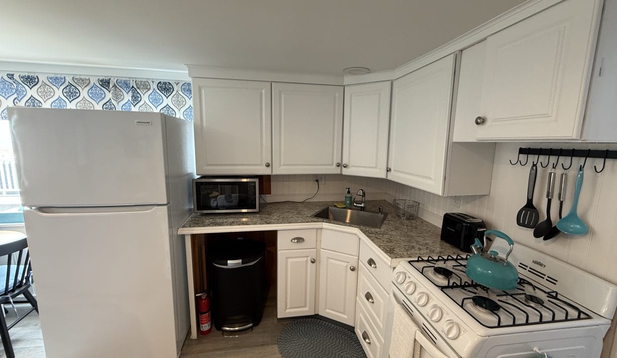 A compact kitchen with white cabinets, a white refrigerator, microwave, gas stove, kettle, utensils, and a sink on marble countertops.