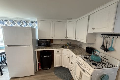 A compact kitchen with white cabinets, a white refrigerator, microwave, gas stove, kettle, utensils, and a sink on marble countertops.