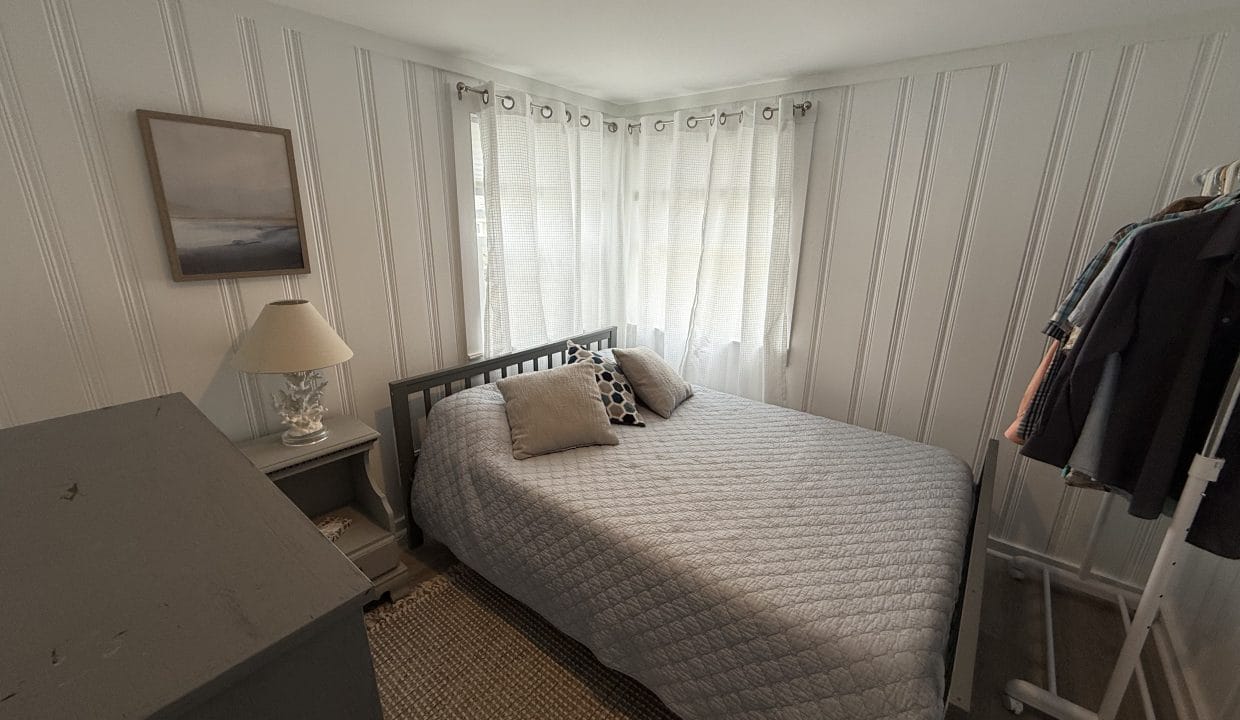 Small bedroom with a gray bed, two pillows, a nightstand with a lamp, a framed picture, a clothes rack with hanging clothes, and white curtains over corner windows.