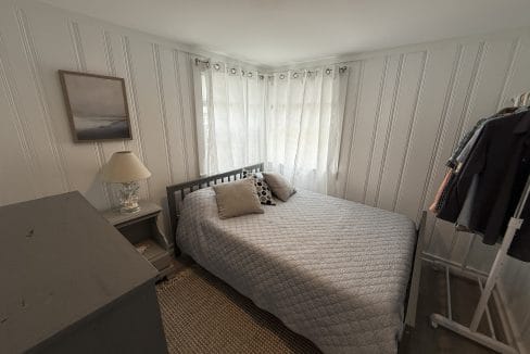 Small bedroom with a gray bed, two pillows, a nightstand with a lamp, a framed picture, a clothes rack with hanging clothes, and white curtains over corner windows.
