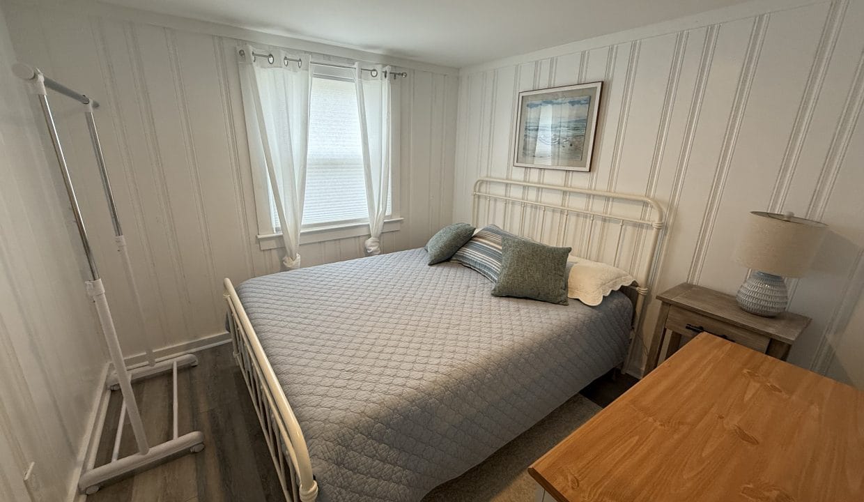 A small bedroom with a metal bed frame, blue quilt, two pillows, a nightstand with a lamp, dresser, wall art, and a garment rack by two curtained windows.