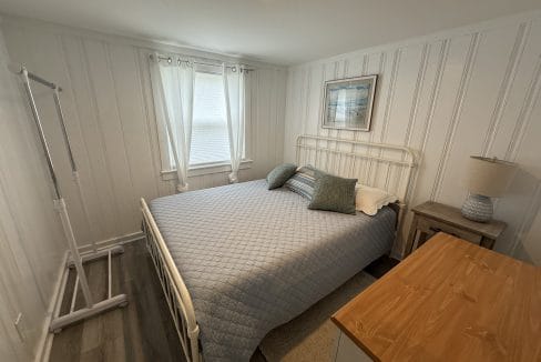 A small bedroom with a metal bed frame, blue quilt, two pillows, a nightstand with a lamp, dresser, wall art, and a garment rack by two curtained windows.
