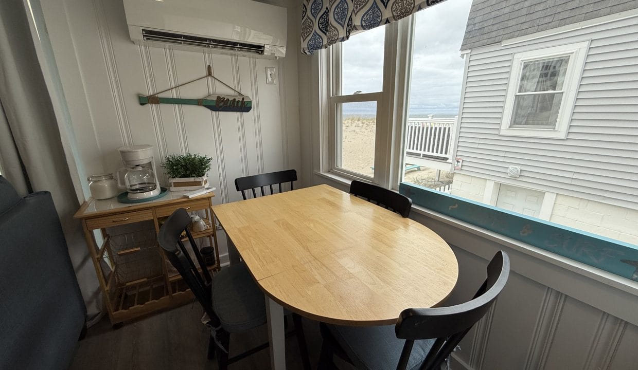 A small dining area with a wooden table and four chairs beside a window, with a coffee station and beach-themed decor on the wall.