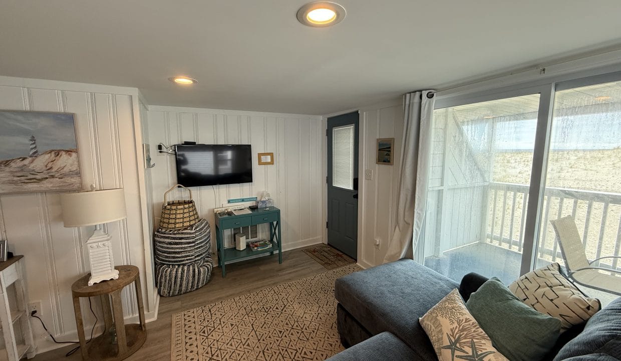 A cozy living room with a blue sectional sofa, wall-mounted TV, decorative baskets, a small table, and sliding glass doors leading to a balcony.