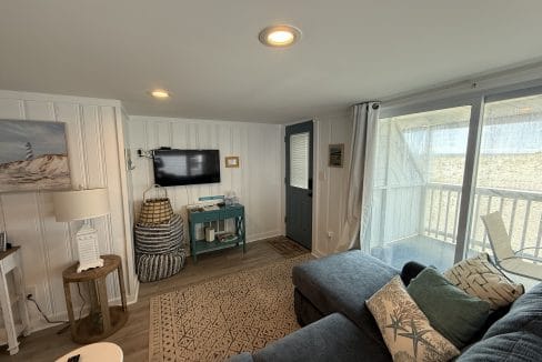 A cozy living room with a blue sectional sofa, wall-mounted TV, decorative baskets, a small table, and sliding glass doors leading to a balcony.