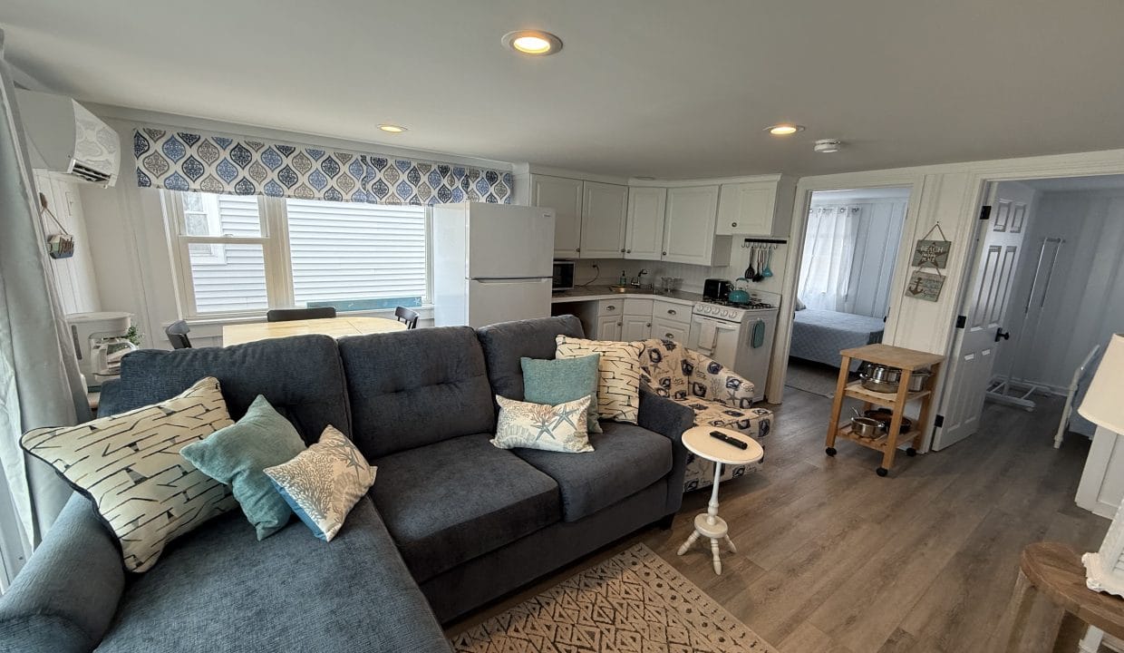 A cozy living room with a blue sectional sofa, patterned armchair, and neutral decor opens to a white kitchen and a bedroom visible through an open door.