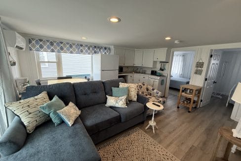 A cozy living room with a blue sectional sofa, patterned armchair, and neutral decor opens to a white kitchen and a bedroom visible through an open door.
