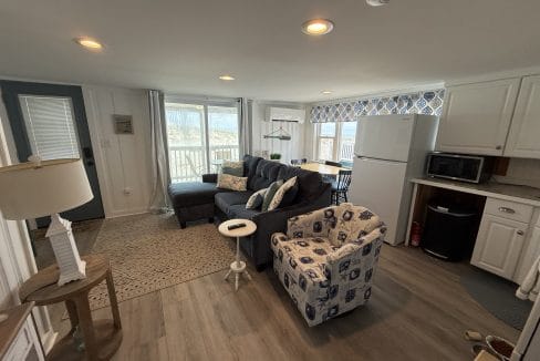 A small, open-plan living area with a patterned armchair, a dark sofa, a dining table, a kitchenette with white cabinets, and large windows overlooking an outdoor deck.