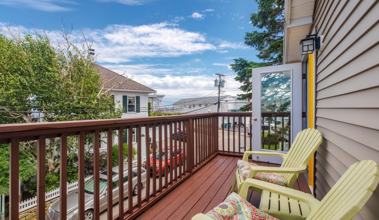 A small wooden balcony with three yellow chairs and a couple of patterned pillows overlooks a residential street with houses, trees, and a clear blue sky in the background.