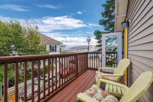 A small wooden balcony with three yellow chairs and a couple of patterned pillows overlooks a residential street with houses, trees, and a clear blue sky in the background.