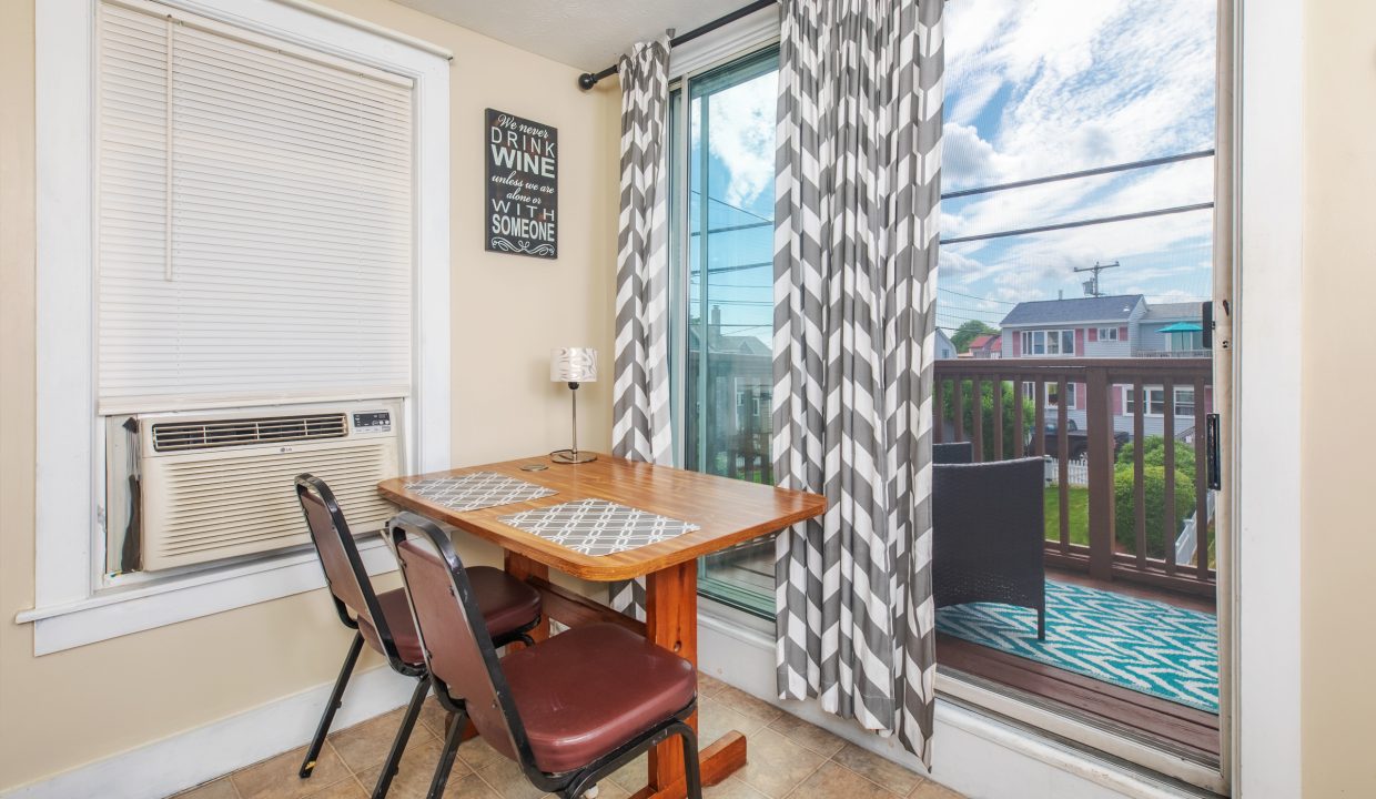 A small dining area with a wooden table, two chairs, a window air conditioner, and sliding glass doors leading to a balcony with outdoor furniture. Curtains and a wall sign are present.