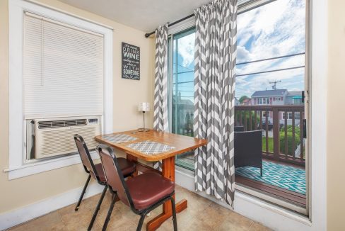 A small dining area with a wooden table, two chairs, a window air conditioner, and sliding glass doors leading to a balcony with outdoor furniture. Curtains and a wall sign are present.