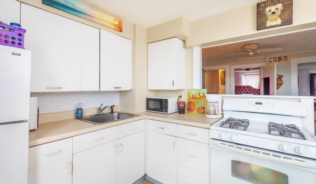 A small kitchen with white cabinets, a gas stove, a sink, a microwave, and a refrigerator. Wall decor includes a colorful painting above cabinets and a 