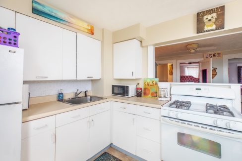 A small kitchen with white cabinets, a gas stove, a sink, a microwave, and a refrigerator. Wall decor includes a colorful painting above cabinets and a 