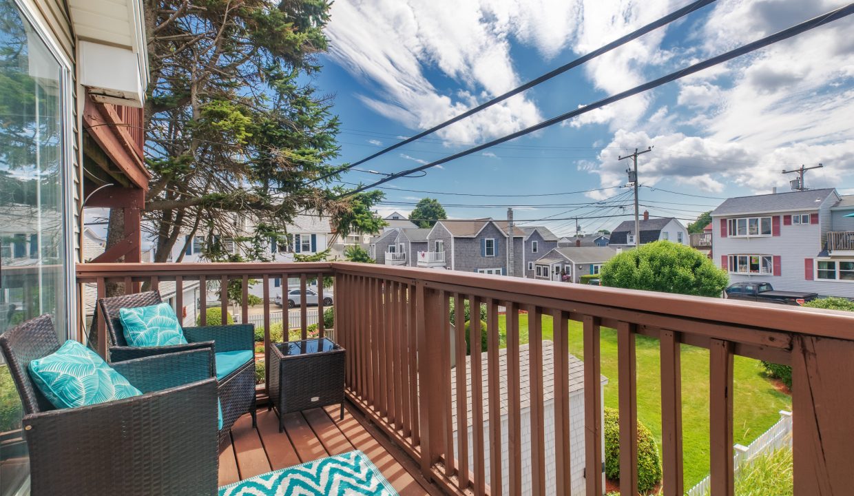 A small balcony with wicker chairs and a table overlooks a suburban neighborhood with houses, trees, and power lines under a partly cloudy sky.