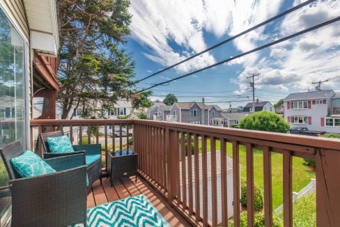 A small balcony with wicker chairs and a table overlooks a suburban neighborhood with houses, trees, and power lines under a partly cloudy sky.