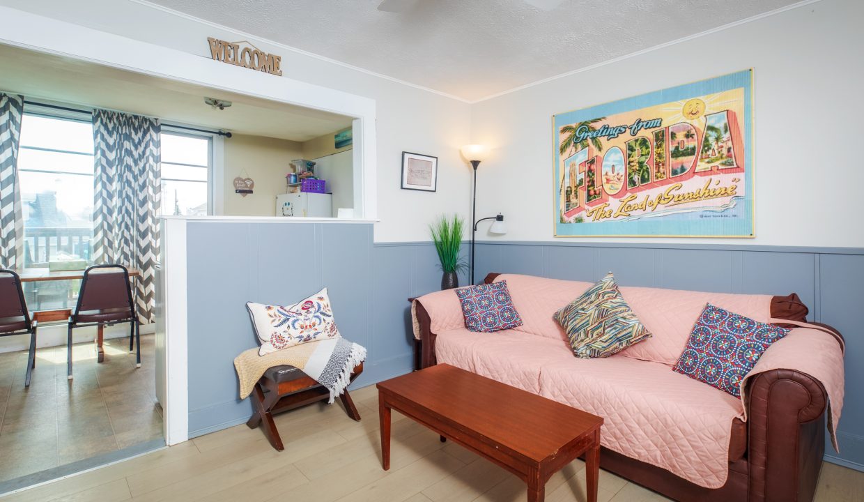 A cozy living room with a brown sofa covered by a pink throw, two patterned pillows, a wooden coffee table, a small chair, and a 'Welcome to Florida' poster on the wall. Adjoining dining area visible.