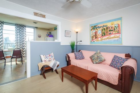 A cozy living room with a brown sofa covered by a pink throw, two patterned pillows, a wooden coffee table, a small chair, and a 'Welcome to Florida' poster on the wall. Adjoining dining area visible.