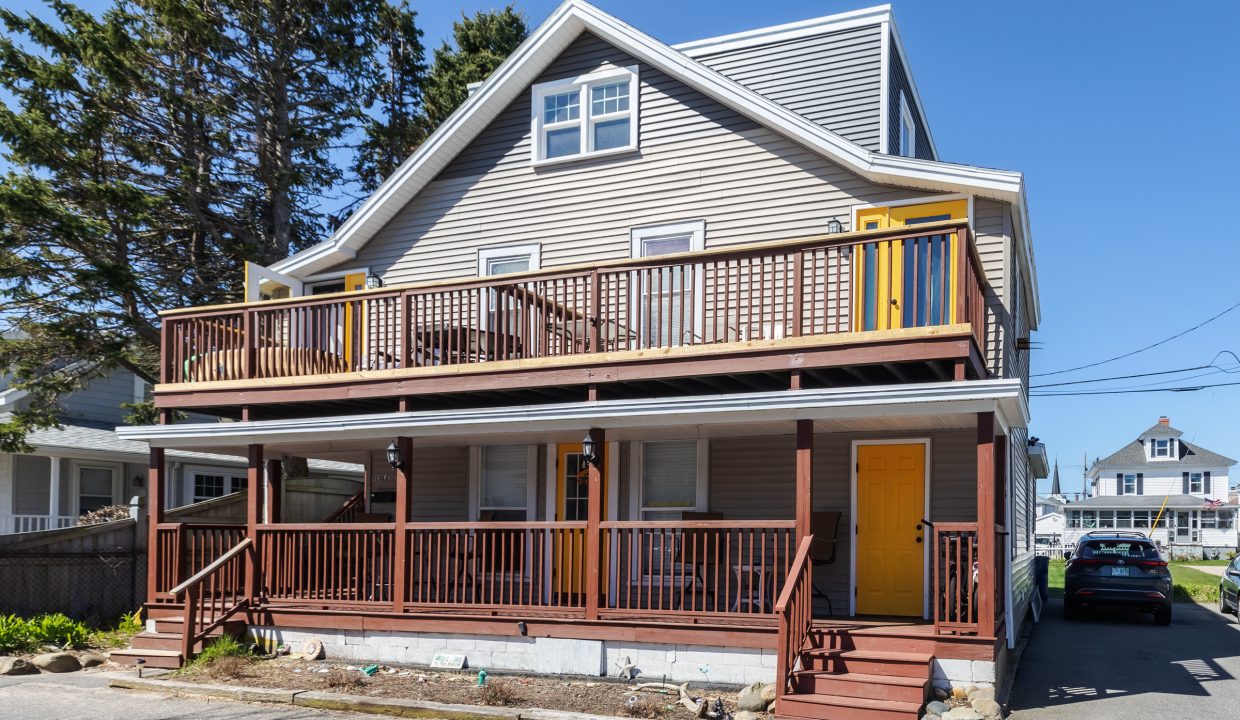 Two-story house with a wrap-around wooden porch, gray siding, and a yellow door, located on a sunny residential street with trees.