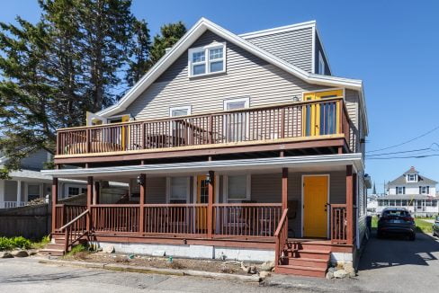 Two-story house with a wrap-around wooden porch, gray siding, and a yellow door, located on a sunny residential street with trees.