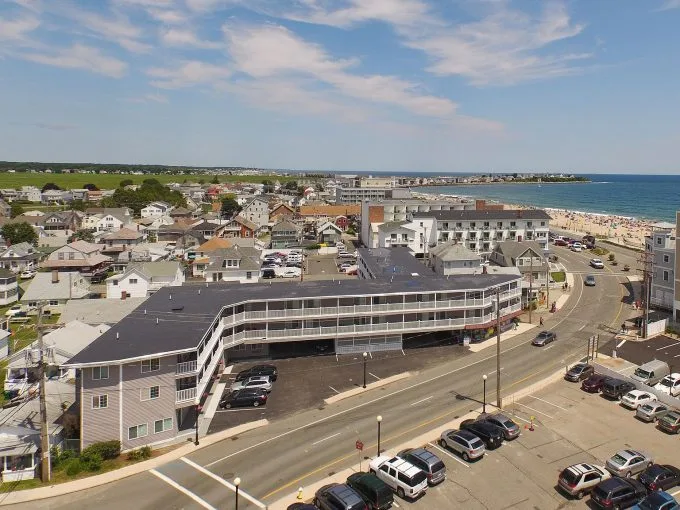 Aerial view of a coastal town with a mix of homes and buildings near the beach, with parked cars along the street and a clear blue sky overhead.