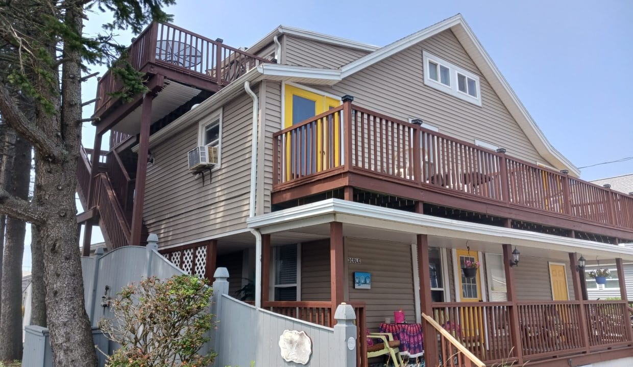 Two-story beige house with brown trim, multiple decks and a yellow door; a person sits on the porch under a tree near a gray fence.