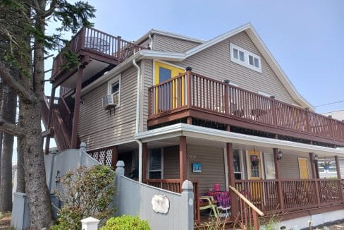 Two-story beige house with brown trim, multiple decks and a yellow door; a person sits on the porch under a tree near a gray fence.