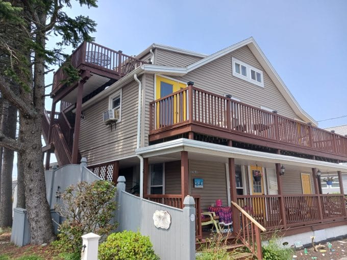 Two-story beige house with brown trim, multiple decks and a yellow door; a person sits on the porch under a tree near a gray fence.