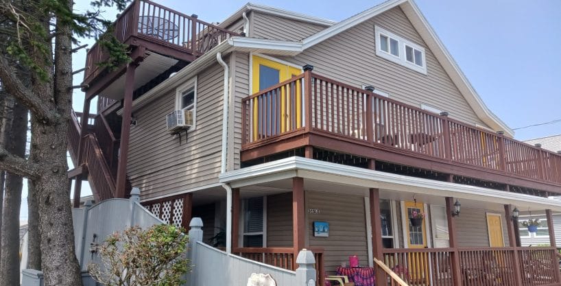 Two-story beige house with brown trim, multiple decks and a yellow door; a person sits on the porch under a tree near a gray fence.