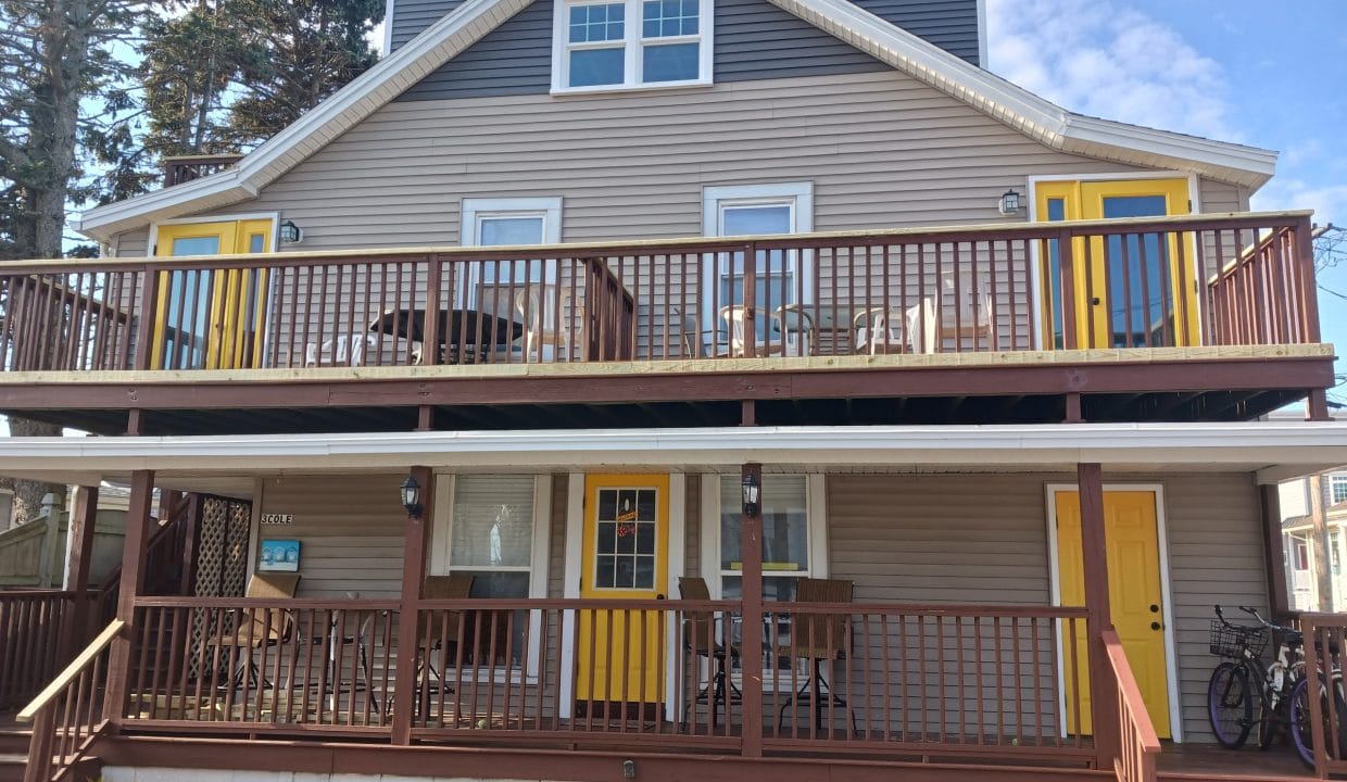 Two-story house with beige siding, brown railings, and three yellow doors. The upper and lower decks have patio furniture, and a bicycle is beside the front stairs.