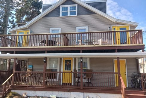 Two-story house with beige siding, brown railings, and three yellow doors. The upper and lower decks have patio furniture, and a bicycle is beside the front stairs.