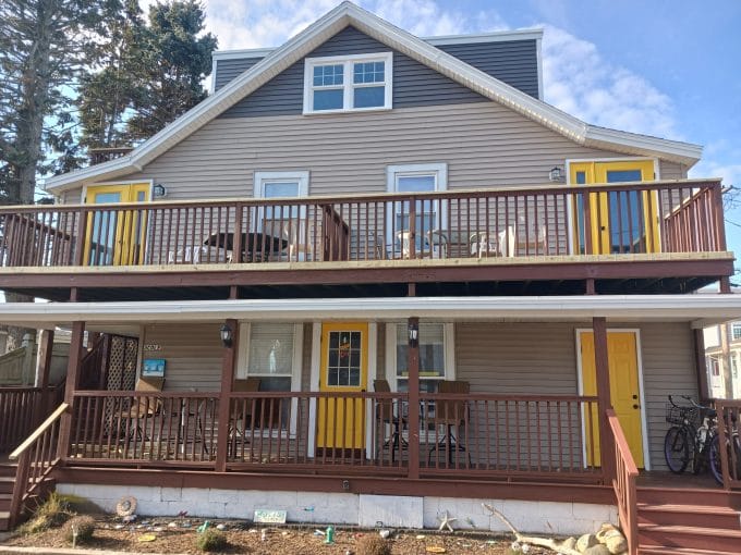 Two-story house with beige siding, brown railings, and three yellow doors. The upper and lower decks have patio furniture, and a bicycle is beside the front stairs.