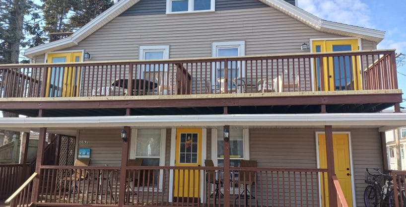 Two-story house with beige siding, brown railings, and three yellow doors. The upper and lower decks have patio furniture, and a bicycle is beside the front stairs.