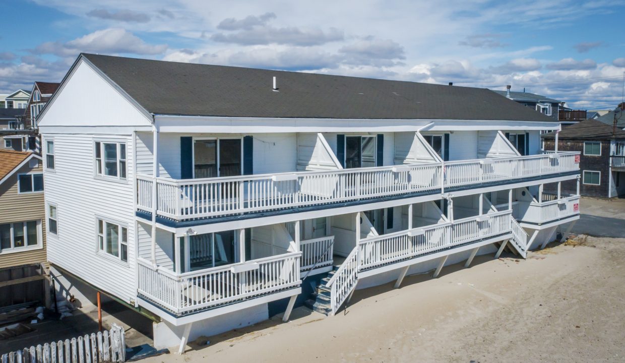 Aerial view of a white multi-story beachfront house with balconies and outdoor stairs, situated on sandy terrain with neighboring houses in the background.