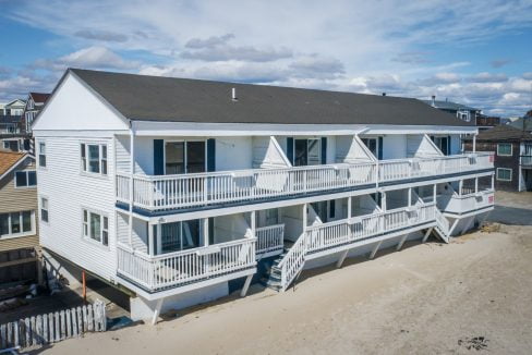 Aerial view of a white multi-story beachfront house with balconies and outdoor stairs, situated on sandy terrain with neighboring houses in the background.