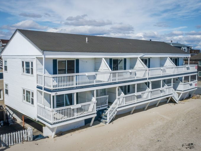 Aerial view of a white multi-story beachfront house with balconies and outdoor stairs, situated on sandy terrain with neighboring houses in the background.