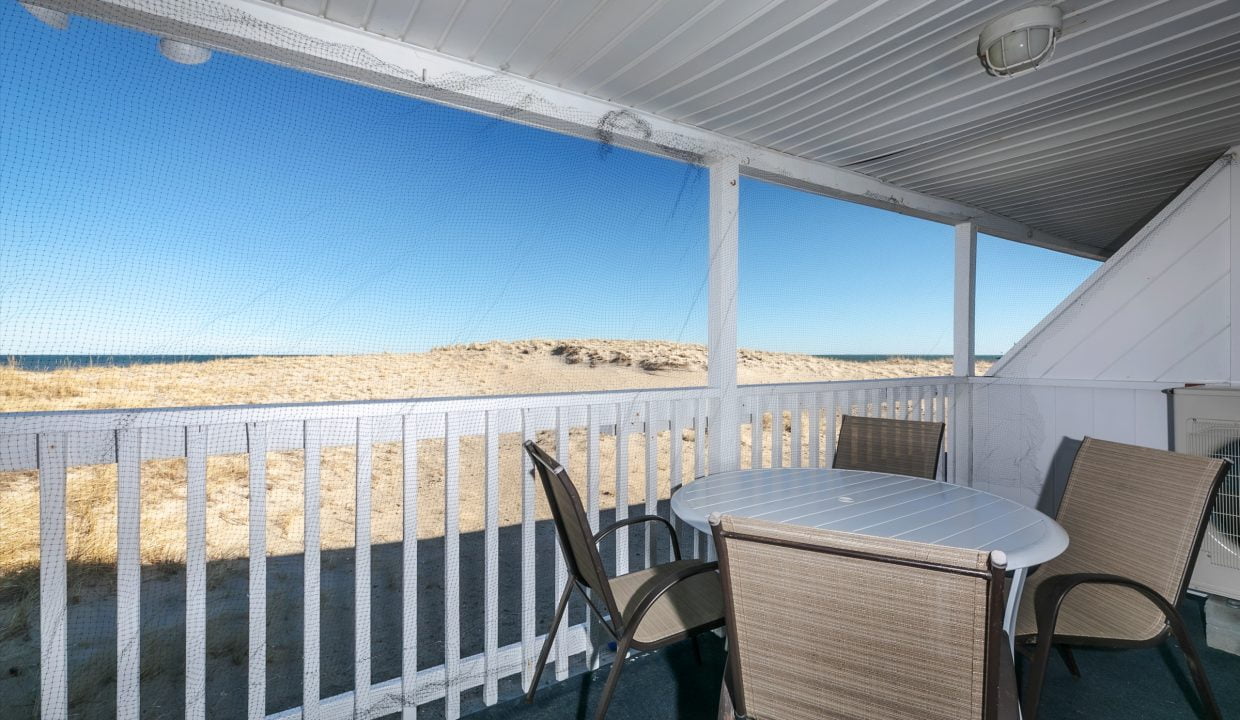 Patio with a table and chairs overlooking a beach dune, enclosed by a screen and white railing.
