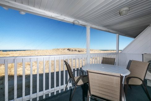 Patio with a table and chairs overlooking a beach dune, enclosed by a screen and white railing.