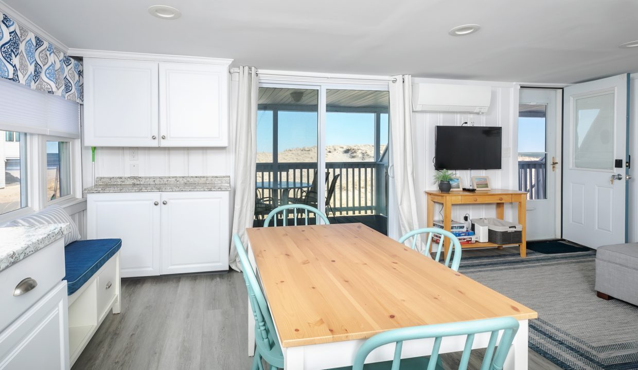 Bright beachfront vacation rental interior with an open-plan living space, wooden dining table, and balcony overlooking the sea.