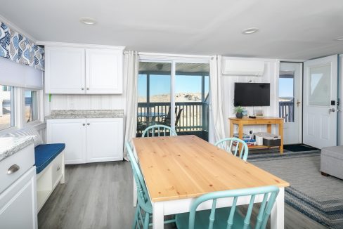 Bright beachfront vacation rental interior with an open-plan living space, wooden dining table, and balcony overlooking the sea.