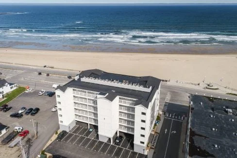 Aerial view of a white multi-story building situated near a beach with a parking lot in front. The ocean and waves are visible in the background, along with scattered parked cars and adjacent buildings.