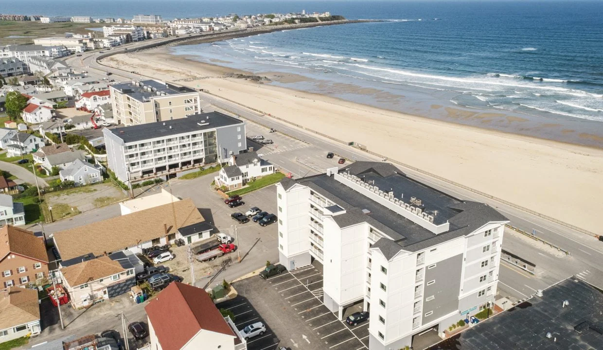 An aerial view of a coastal town with several multi-story buildings near a sandy beach and ocean. The shoreline is visible along with roads and parked cars in the foreground.