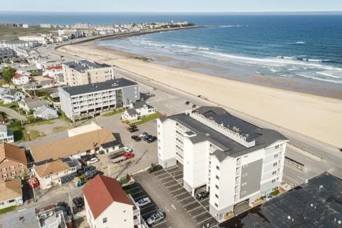 An aerial view of a coastal town with several multi-story buildings near a sandy beach and ocean. The shoreline is visible along with roads and parked cars in the foreground.