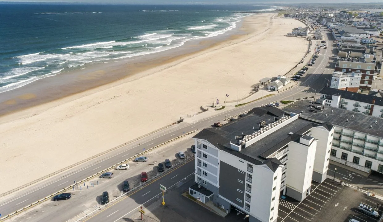 Aerial view of a coastal town featuring a sandy beach, ocean waves, and a road with parked cars parallel to the shoreline. Several buildings are visible along the road.