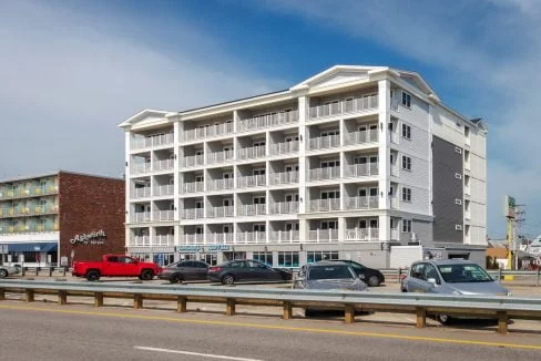 A four-story beachfront hotel with balconies in front of a street with parked cars. A restaurant named 'Angler's' is adjacent on the left. The sky is clear.