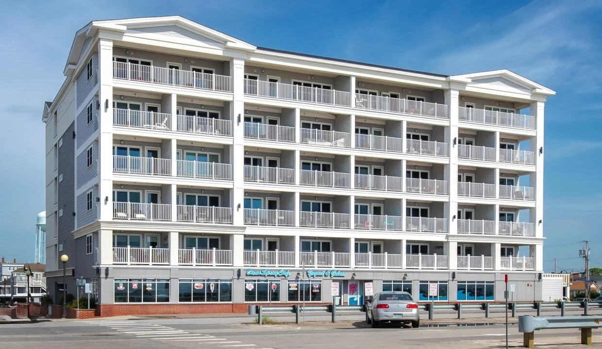 A modern, multi-story beachside building with numerous balconies; storefronts on the ground level; parked cars in front; bright sunny day with a clear blue sky.